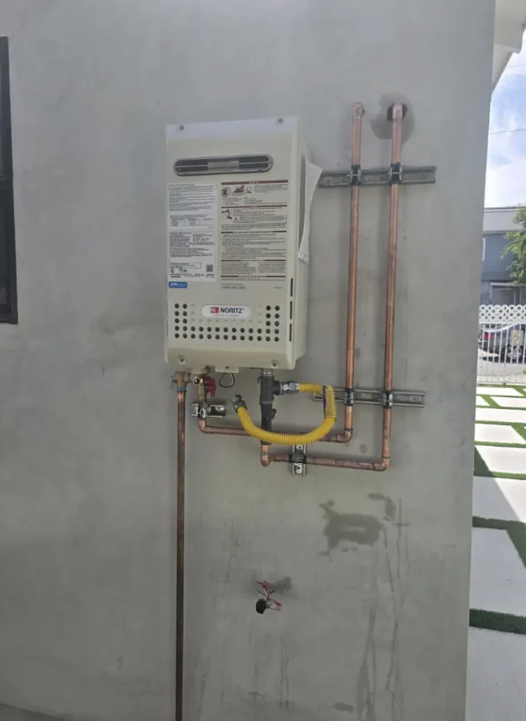 Noritz gas tankless water heater mounted on an exterior stucco wall with copper supply lines and a flexible gas connector